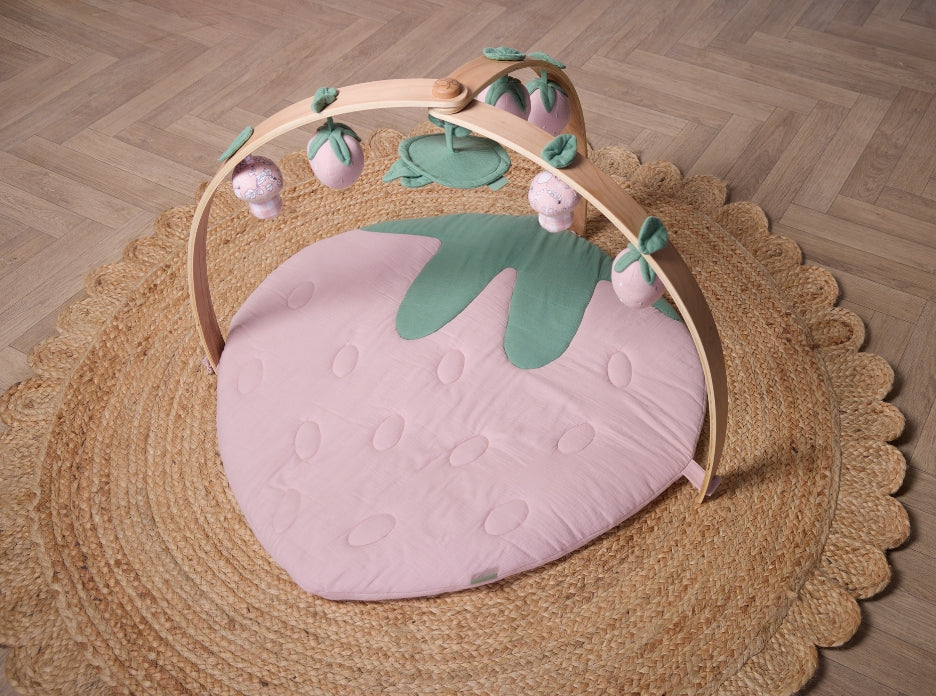 Strawberry-shaped muslin padded playmat with wooden arch and hanging toys styled on woven jute rug.