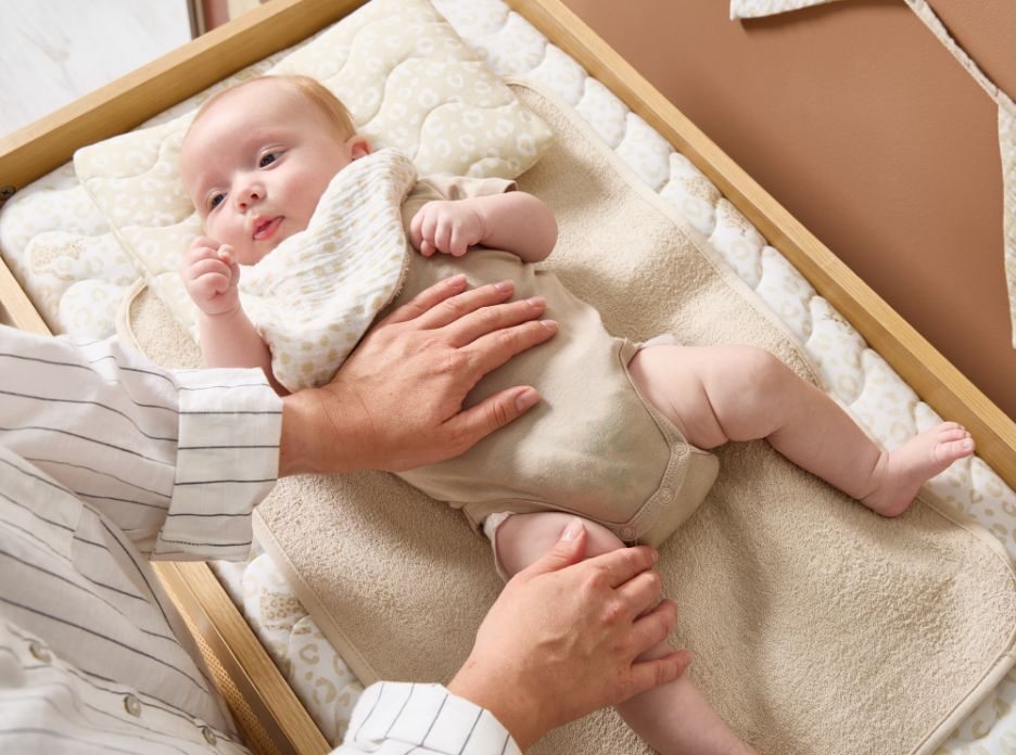 Baby lying on padded changing mat with neutral leopard print cover while parent gently supports during nappy change.