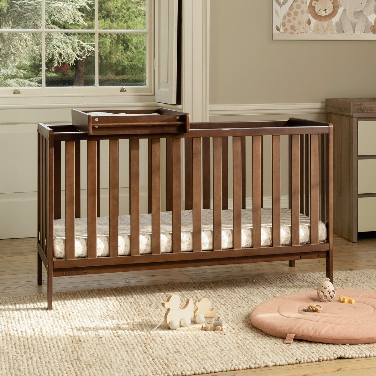 A dark brown wooden convertible cot bed with a matching cot top changer, shown in a styled nursery setting with a beige rug, a pink round floor cushion, and a window in the background.