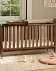 A dark brown wooden convertible cot bed with a matching cot top changer, shown in a styled nursery setting with a beige rug, a pink round floor cushion, and a window in the background.