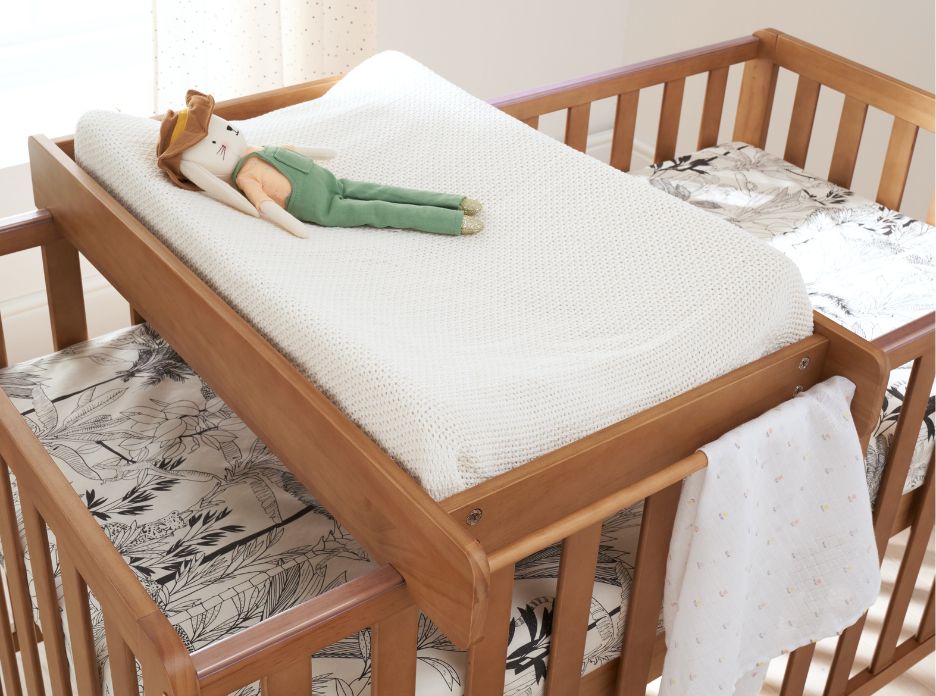 Cot top changer with white changing mat and a teddy on top of a wooden cot