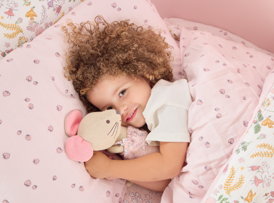 Child lying in bed with Margot Mouse Knitted toy, surrounded by pink and white bedding with floral patterns.