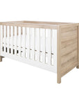 Angled studio view of the Modena Cot Bed in White and Oak featuring clean lines, slatted sides, and a neutral wood finish for a contemporary nursery