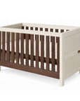 An angle view of the Modena cot bed in cashmere and rich walnut featuring clean lines, slatted sides, and a neutral wood finish for a contemporary nursery.