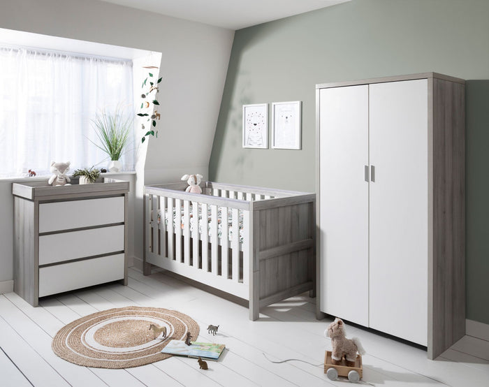 A grey ash and white nursery furniture set including a cot bed, a chest of drawers with a changer, and a wardrobe in a room with light decor.