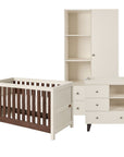 A three-piece nursery furniture set in warm off-white and dark brown wood, including a convertible cot bed, a changing table, and a tall wardrobe, shown on a solid white background.