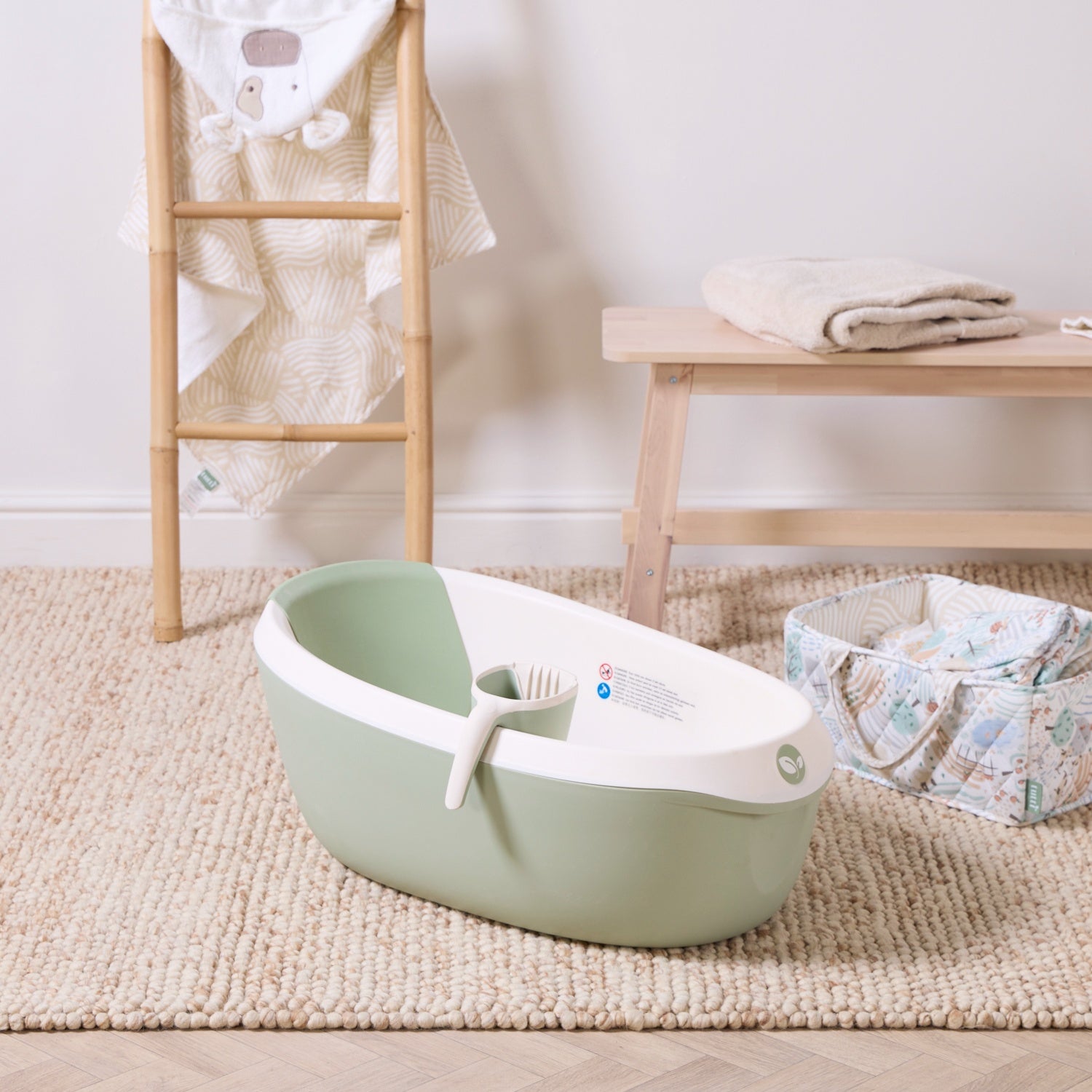 Nati Baby Bath in aloe with matching jug, placed on a textured rug beside soft nursery towels and nappy caddy.