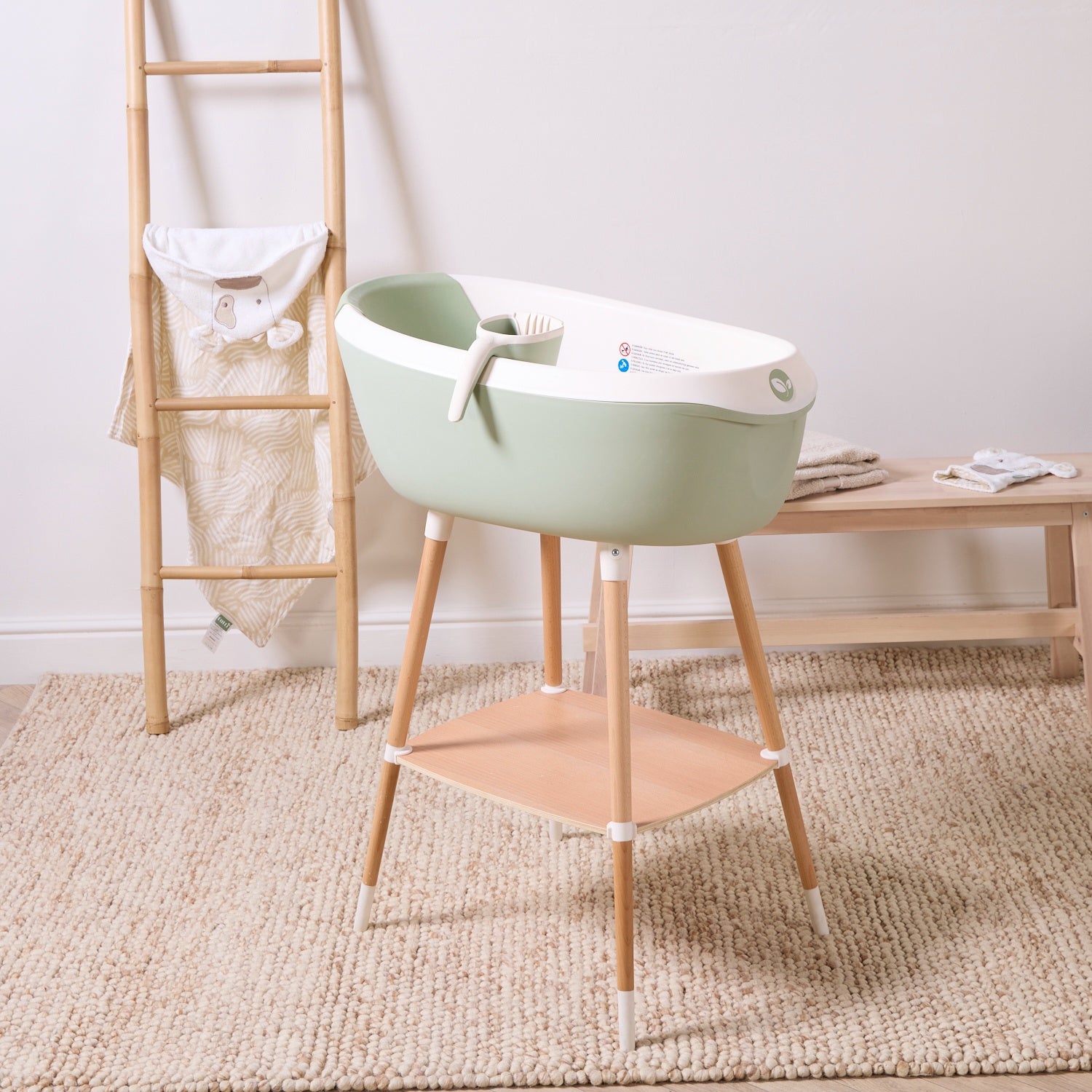 Nati Baby Bath in aloe on wooden stand with matching jug and storage shelf, styled in a calm neutral nursery setting.