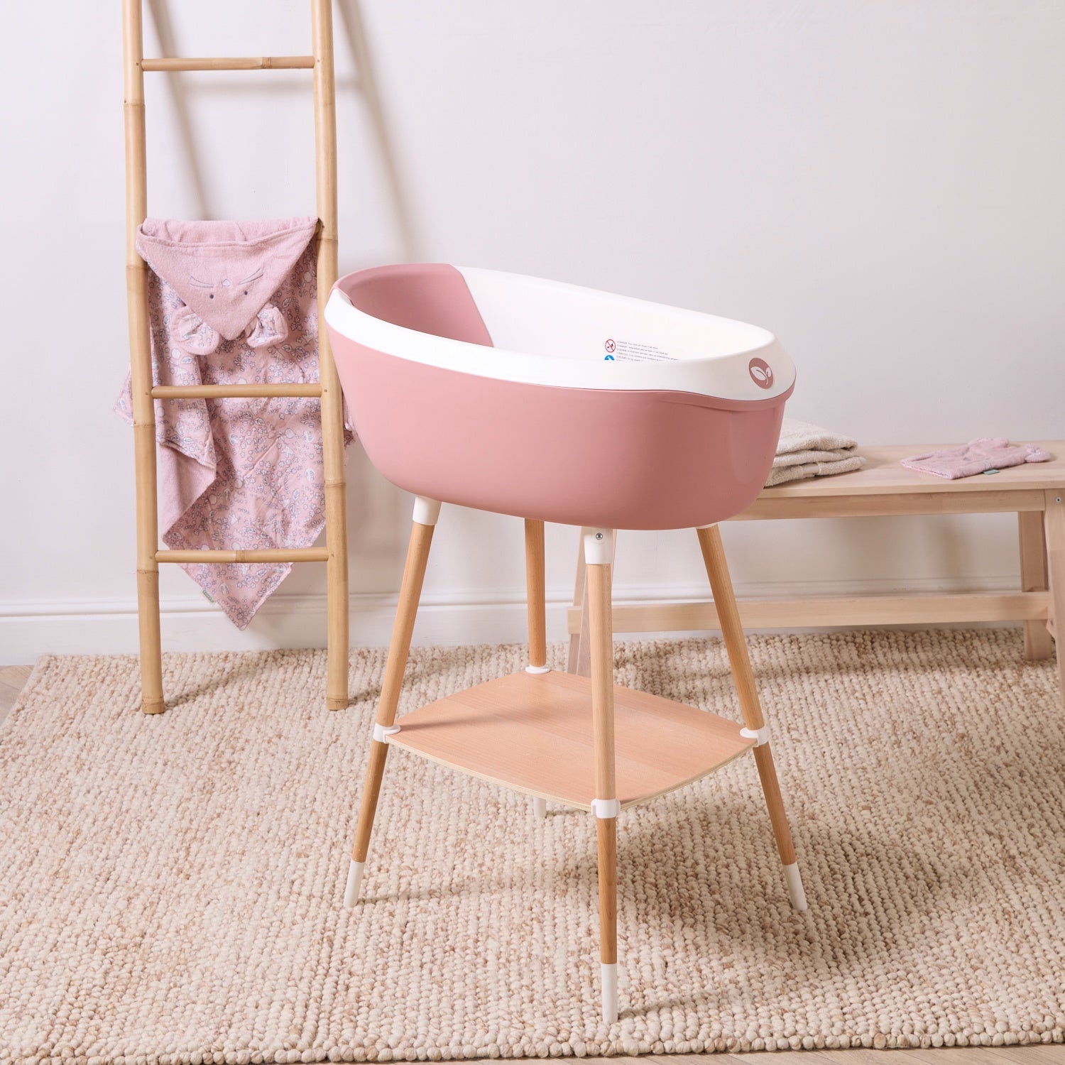 Nati Baby Bath in shell on wooden stand with integrated shelf, styled in a soft nursery space with towels and accessories.