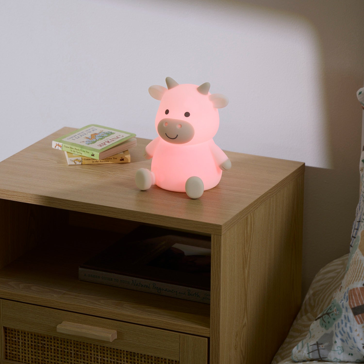Pink cow-shaped night light on a wooden nightstand with a book next to it.