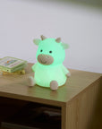 Glowing green cow-shaped night light on a wooden surface with books in the background.