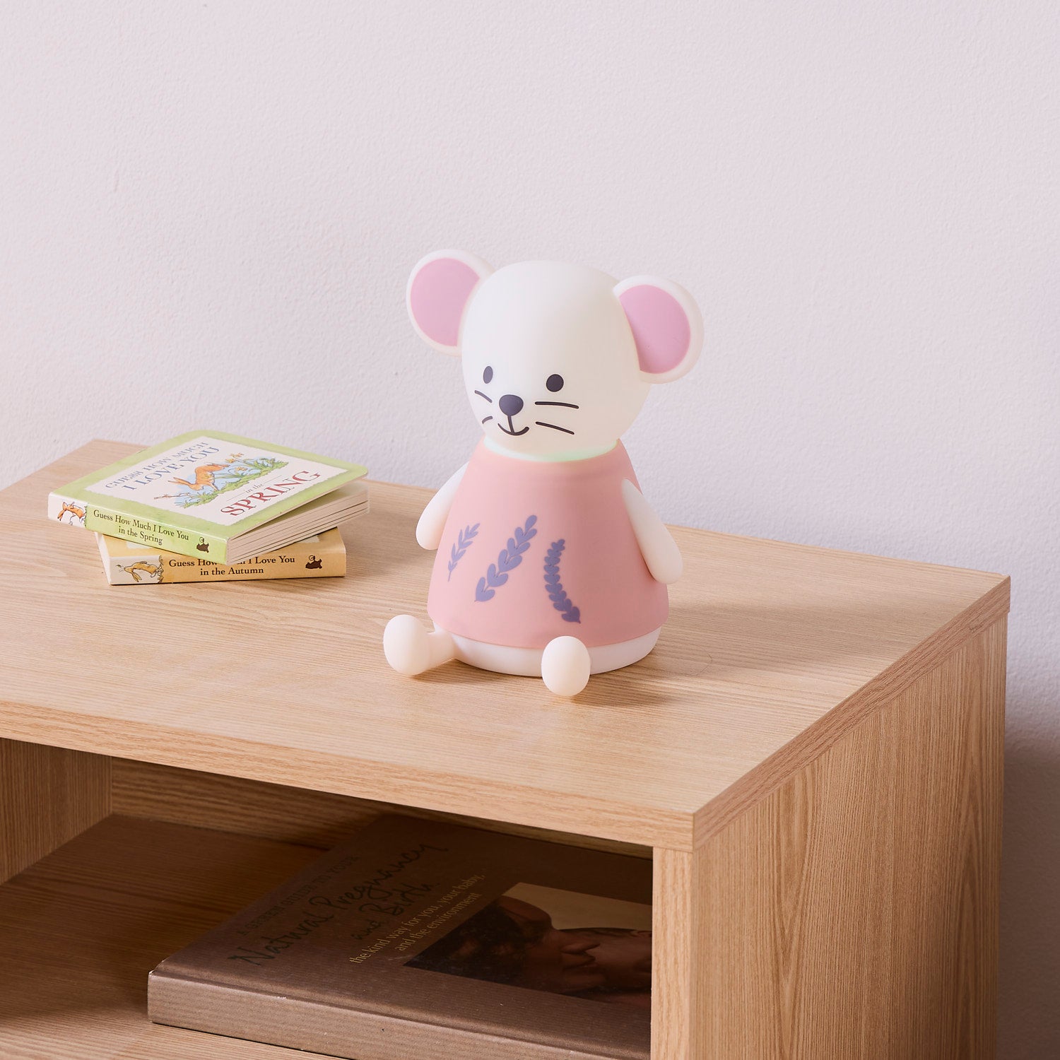 Mouse night light in white with a pink dress on a wooden nightstand with books below.
