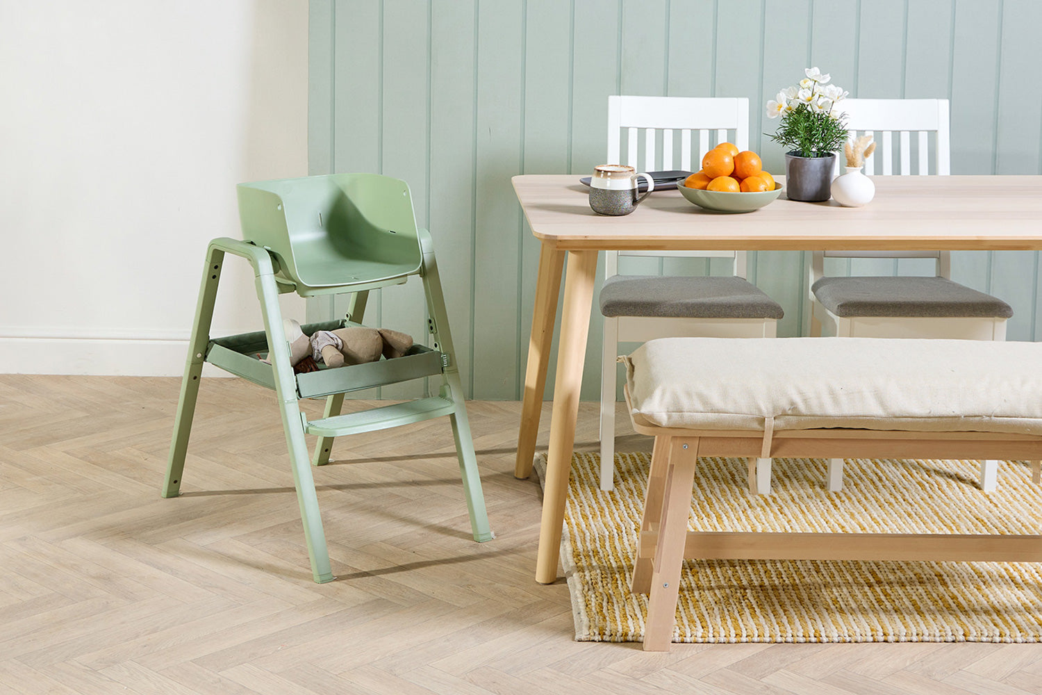 Green highchair next to a wooden dining table with a plant and fruit on a light wood floor.