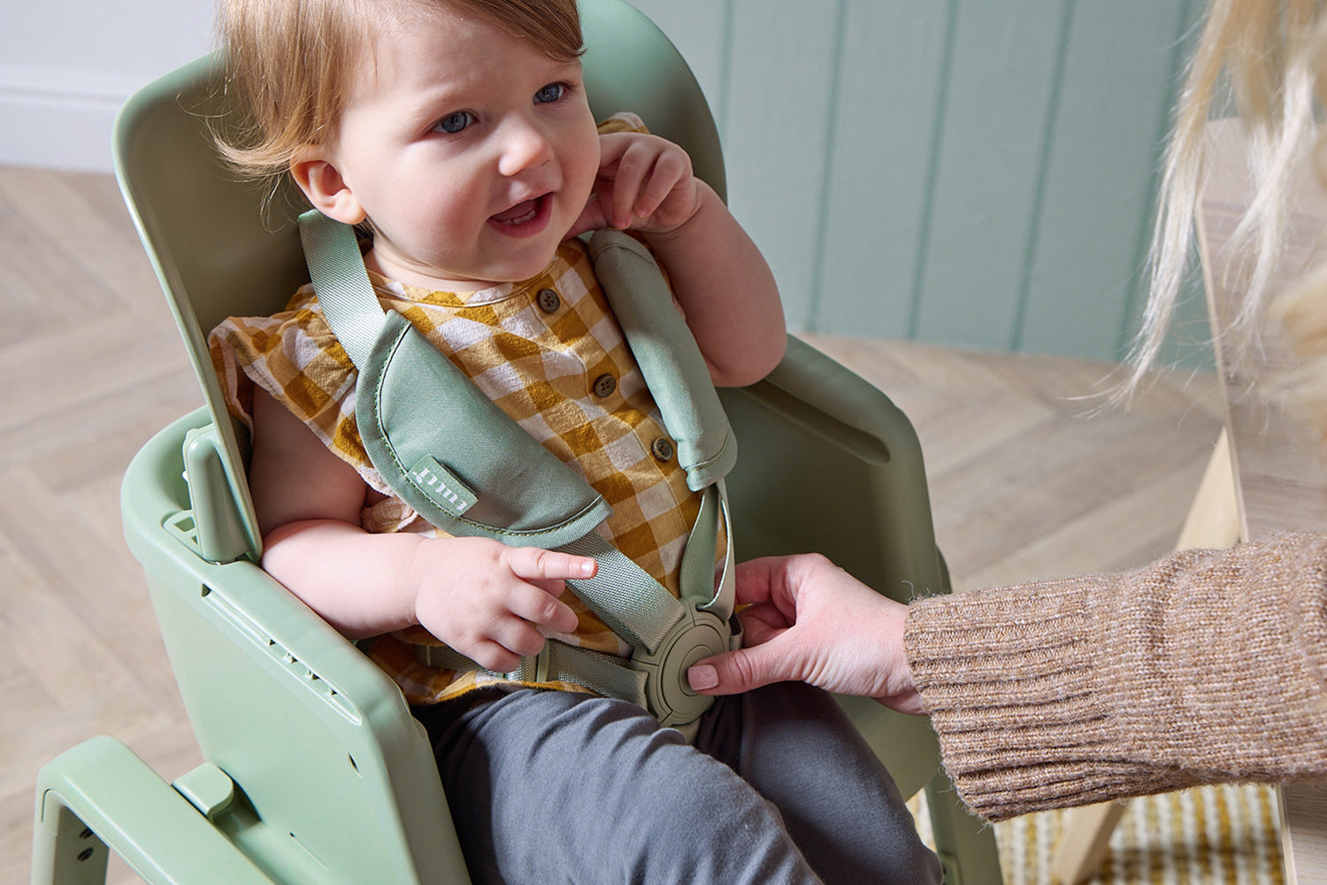 Child in the Nova green highchair with a checkered bib, being adjusted by an adult.