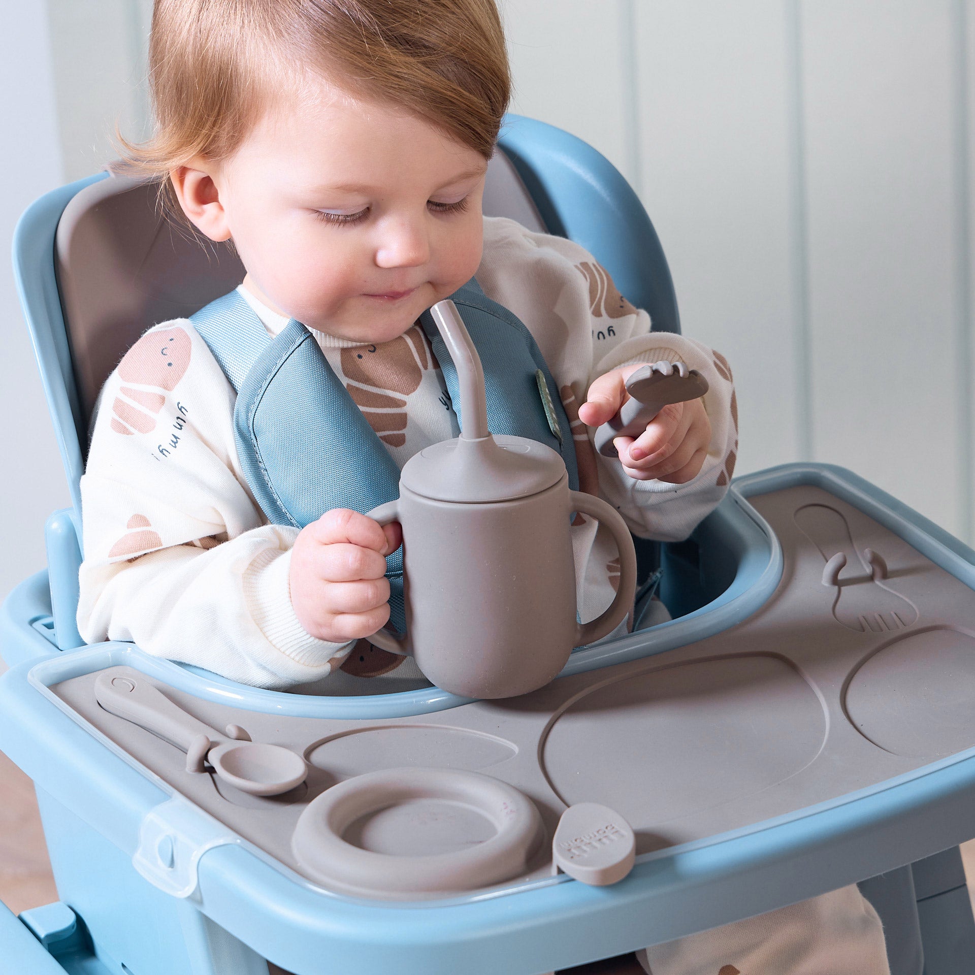 A child sitting in the Nova Maison highchair and holding the sippy cup and fork of the accessory pack.