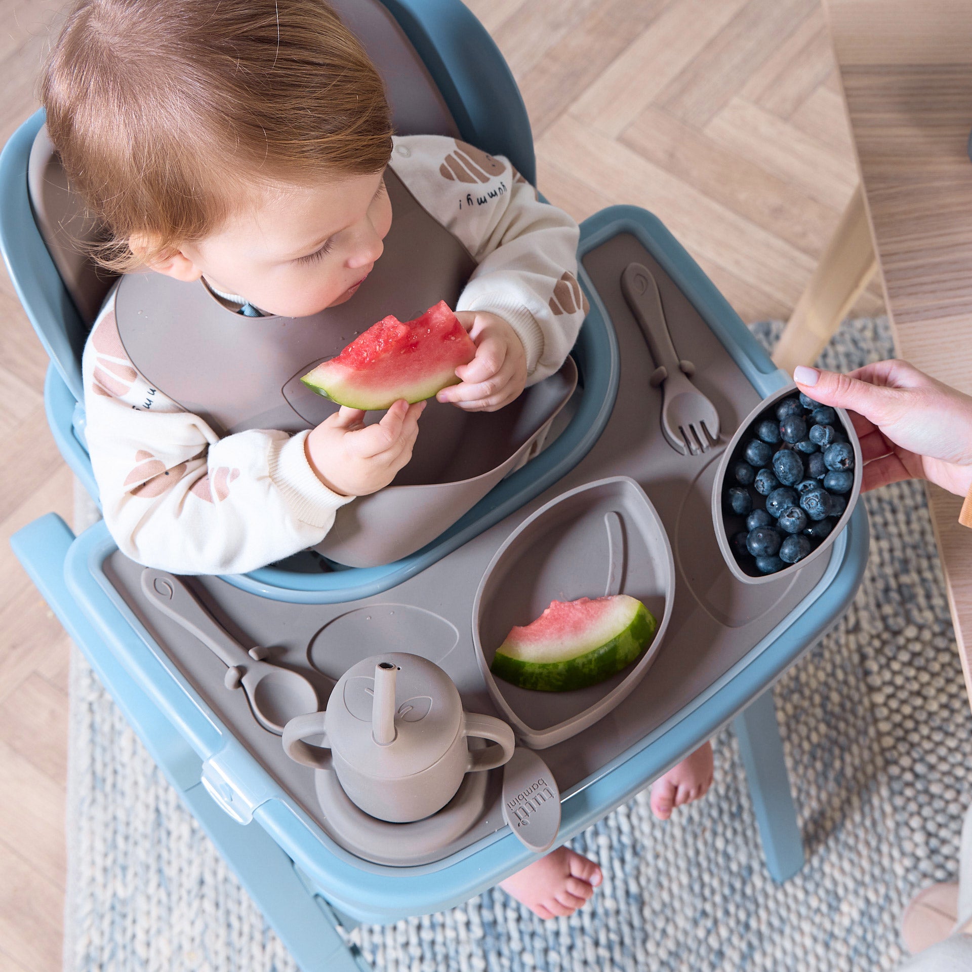 A child eating fruit sitting on the Nova Maison highchair, using the accessory pack, which includes a tray, sippy cup, three nesting bowls, fork, spoon and bib.