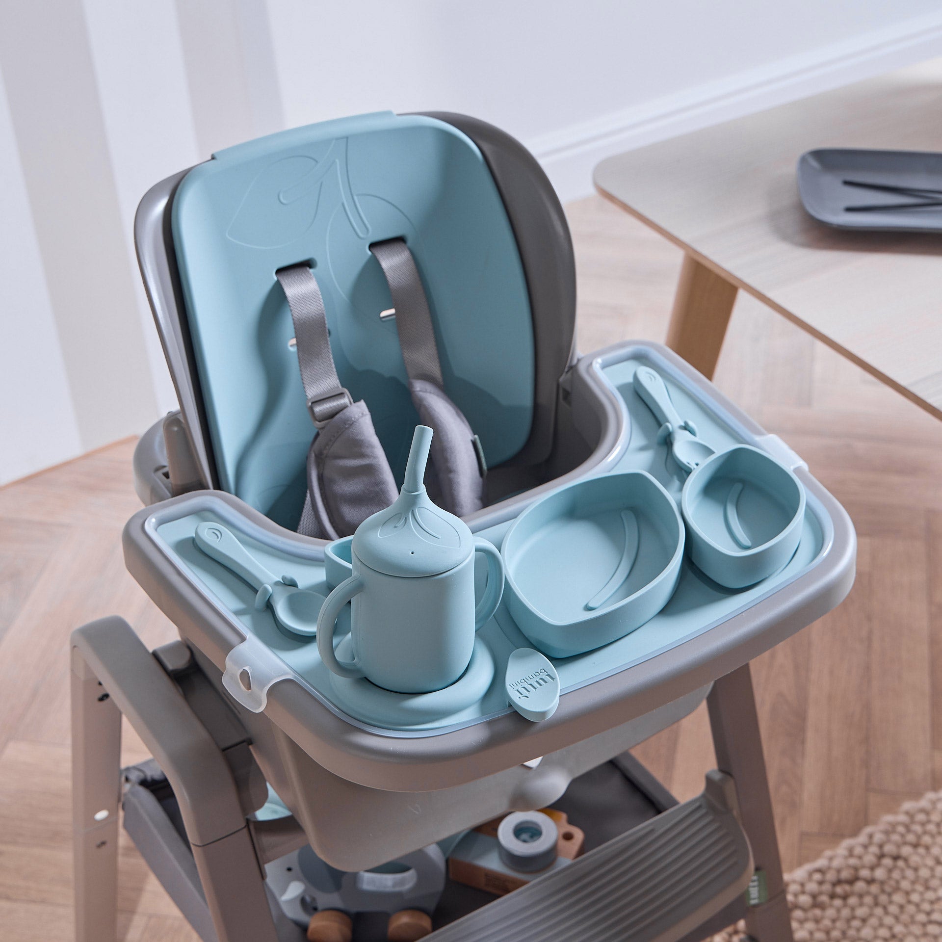 Nova Maison highchair with the accessory pack in blue, including a tray, sippy cup, three nesting bowls, fork and spoon, on a wooden floor.