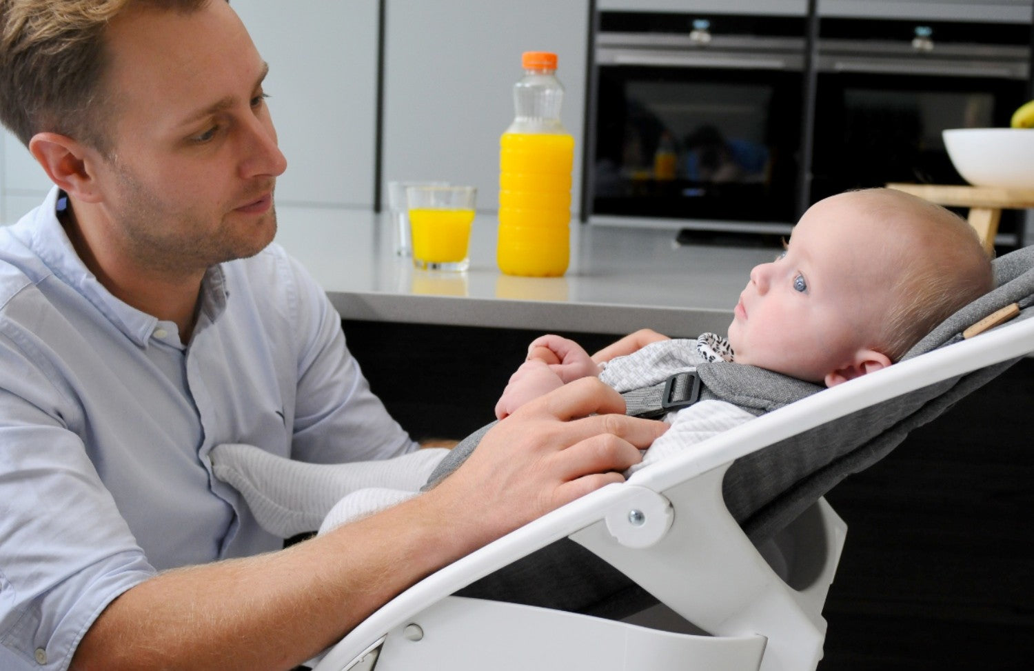 Father gently interacts with his baby seated in the Nova Rocker, showing secure harness comfort and a close bonding moment at home