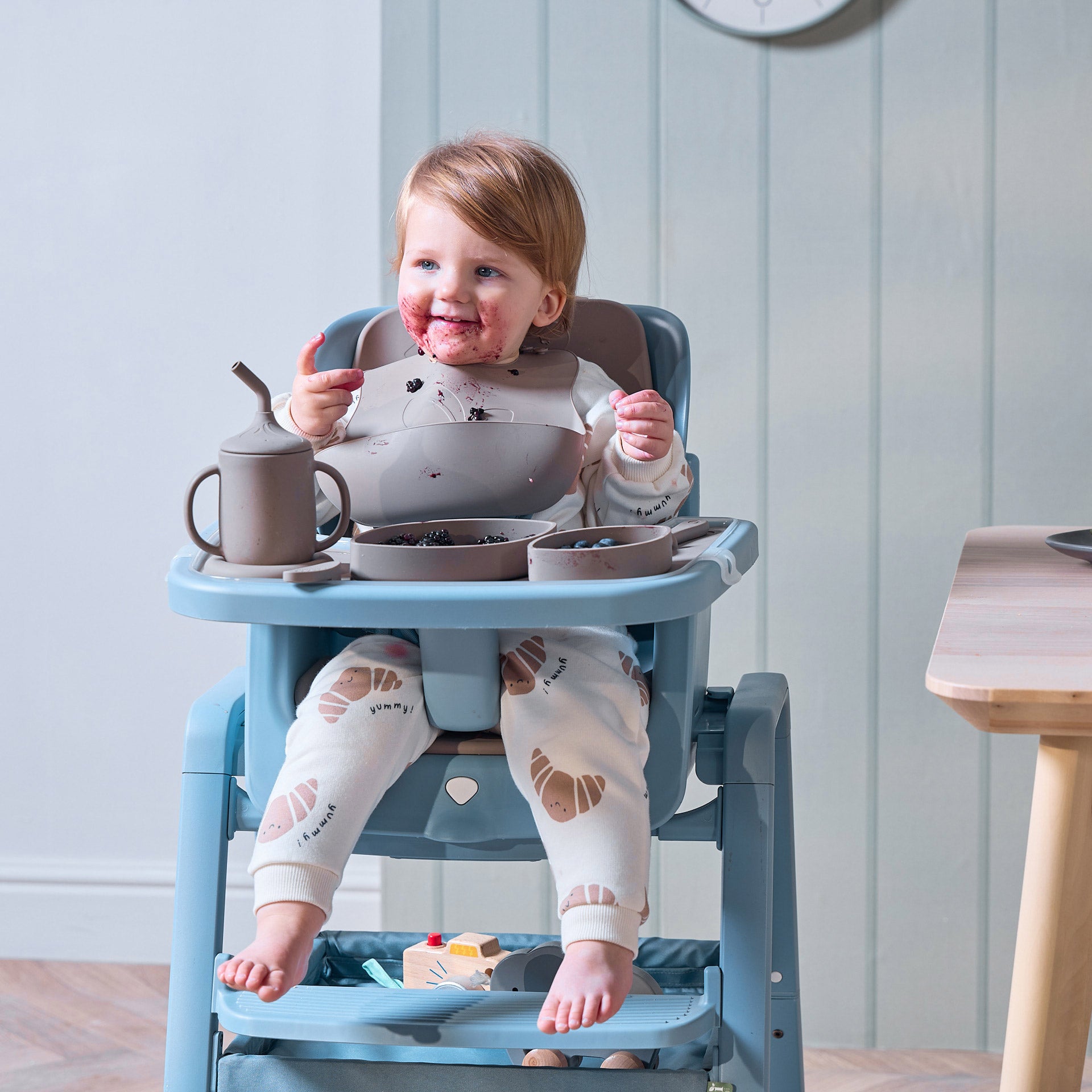 A child eating fruit sitting on the Nova Maison highchair, using the accessory pack that includes a sippy cup, nesting bowls, fork and spoon. 