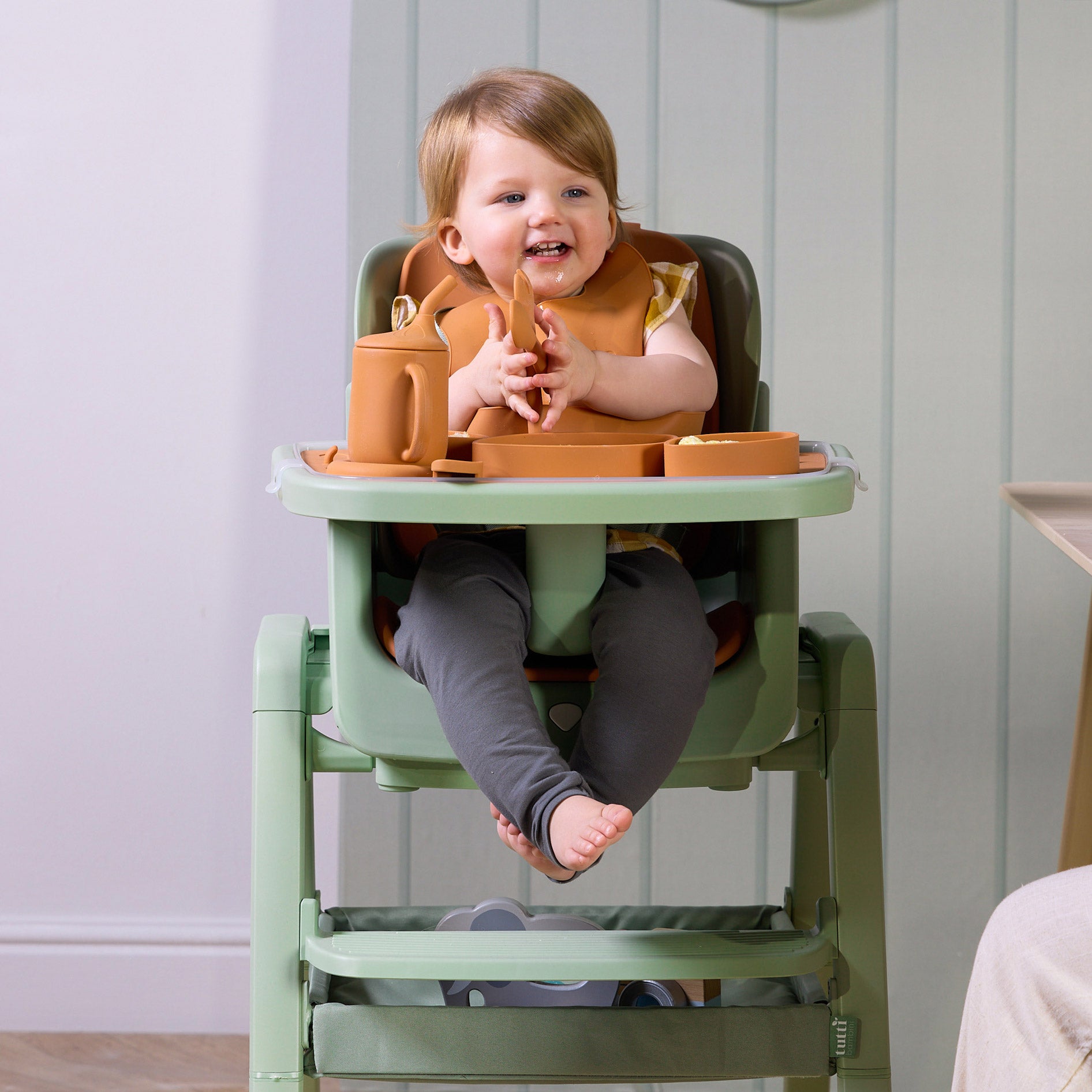 A child in the Nova Maison highchair in green with orange silicone utensils.