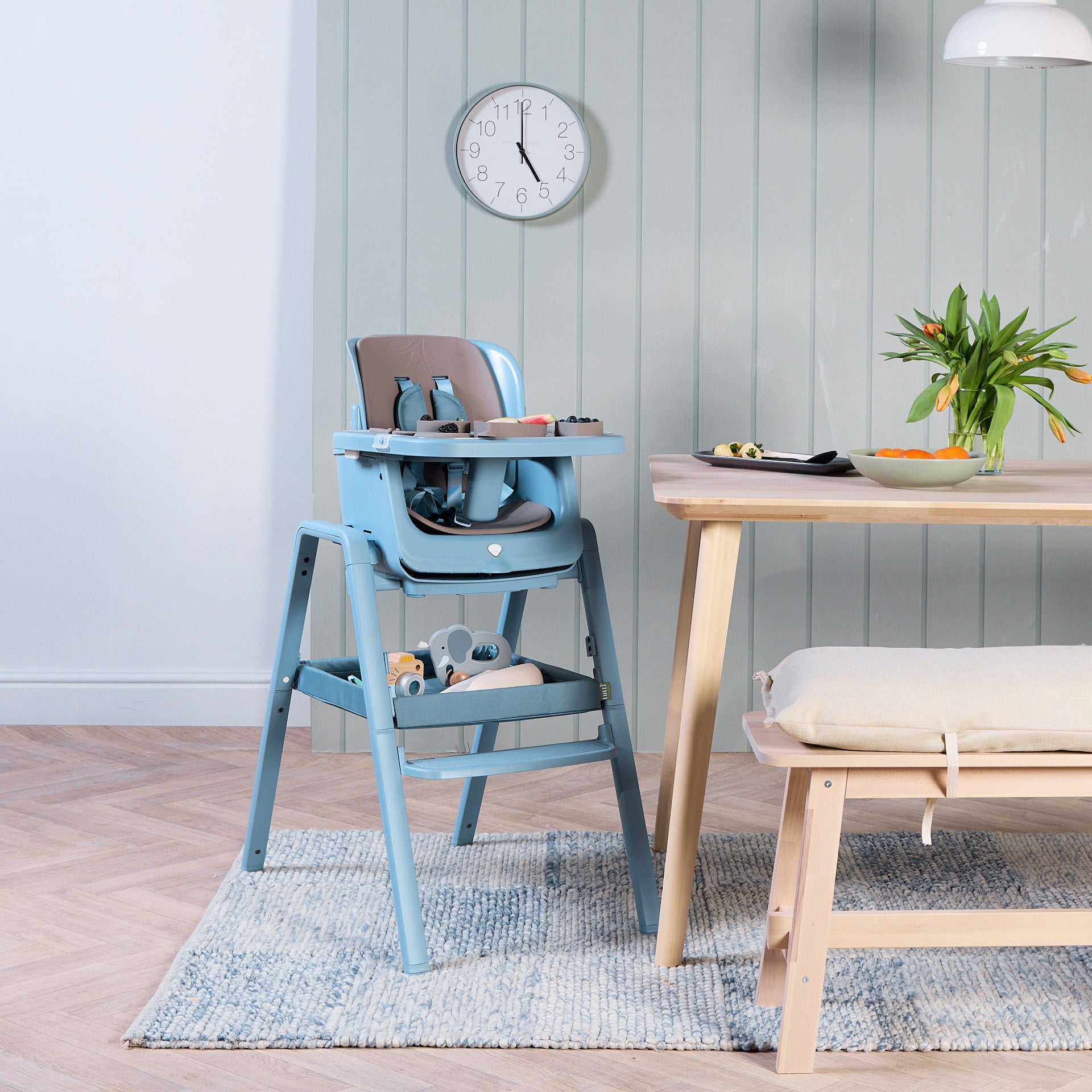 Nova Maison 7 in 1 highchair in blue with the accessory pack in a dinning setting with a table and chair and a clock on the wall.
