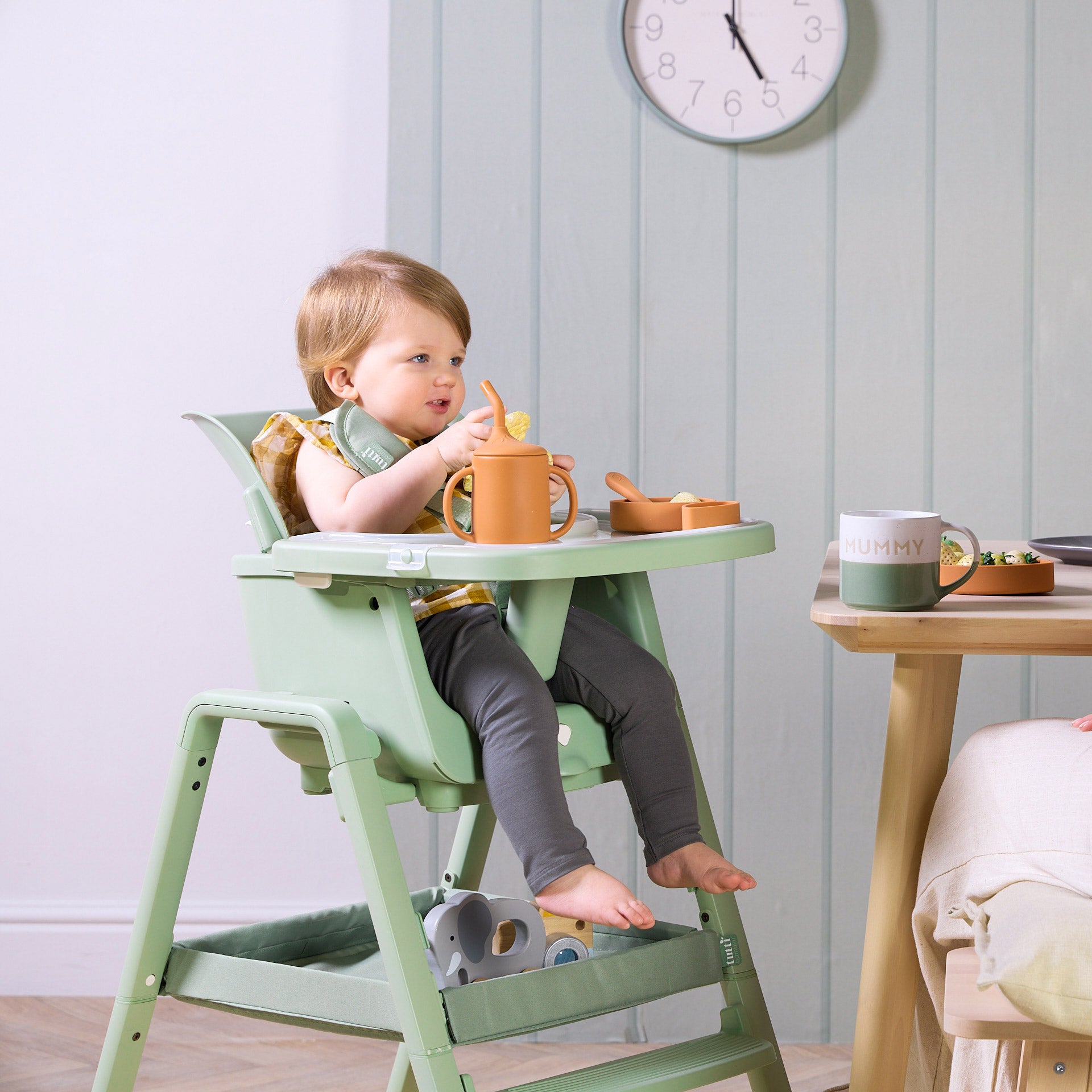 A child sitting in the green Nova Maison high chair with a sippy cup and bowl, in a room with a clock on the wall.