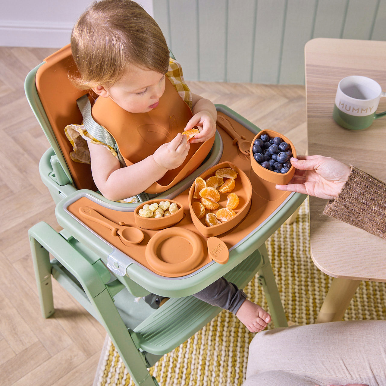 A child in the Nova Maison highchair with a tray, eating fruits, wearing an orange bib.