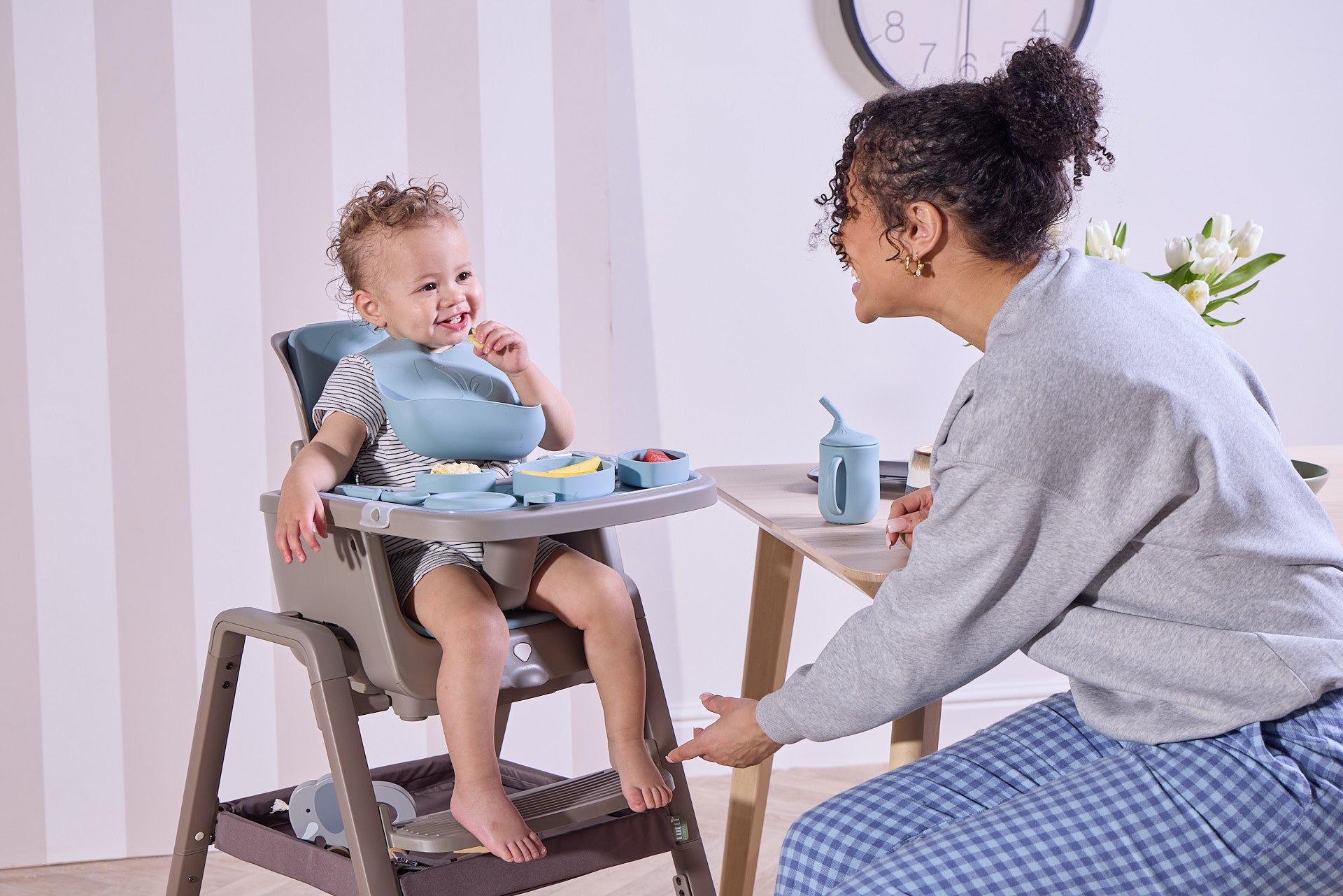 Child in a high chair with a woman sitting next to them, both engaged in mealtime.