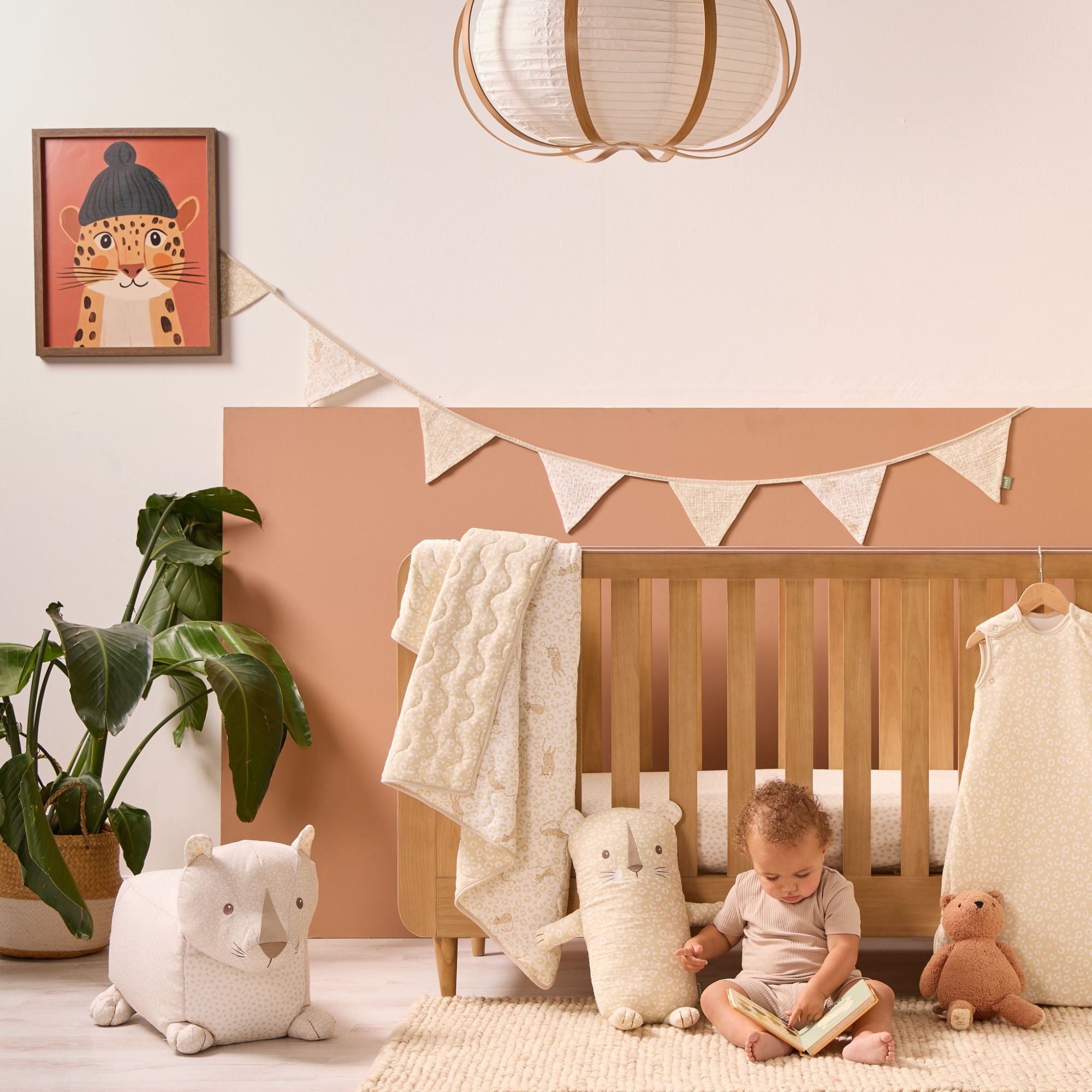 Wooden cot styled with neutral leopard print bedding and love cats nursery bunting as baby sits reading on rug beside soft toy cushions.