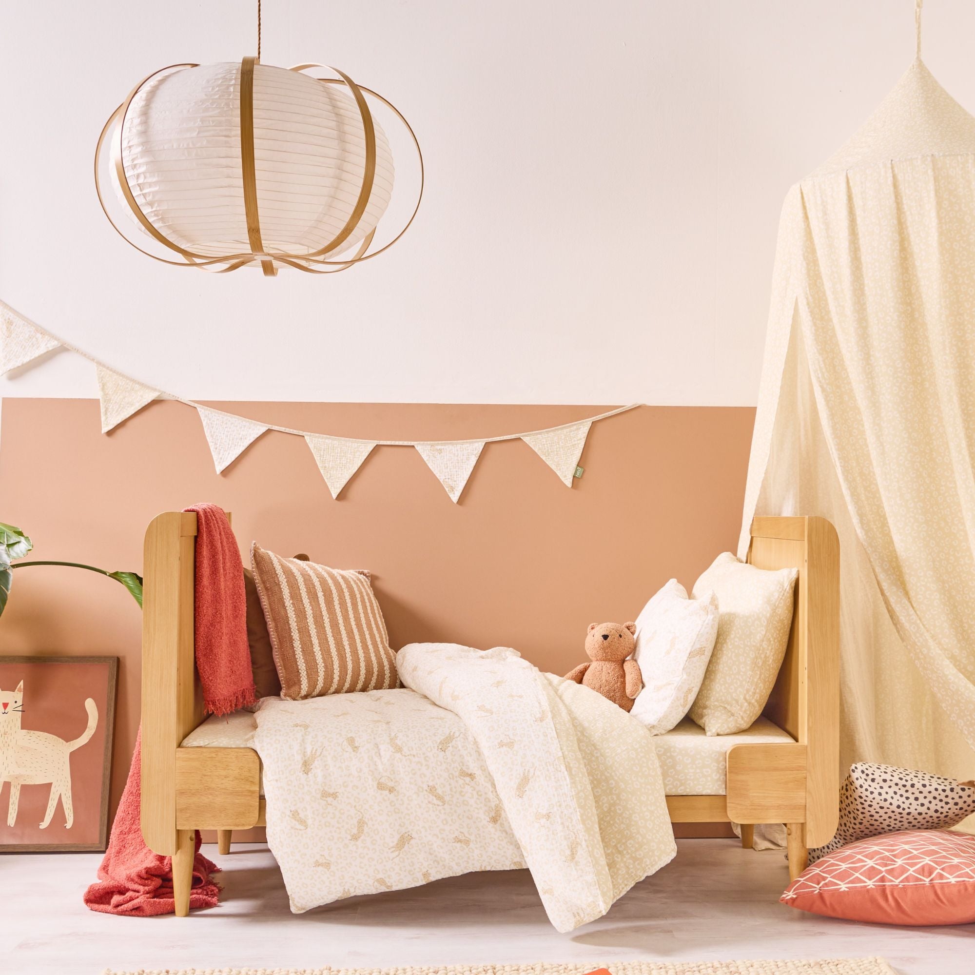 Neutral nursery styled with wooden toddler bed, cream patterned bedding, coordinating bunting and soft canopy in warm tones.