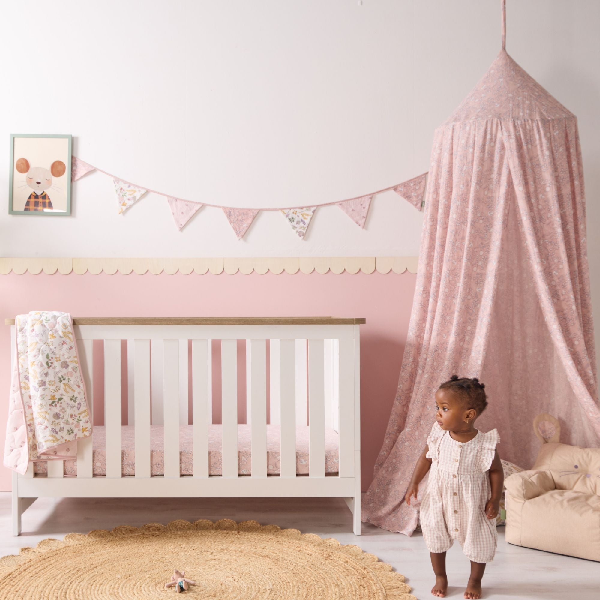 Baby girl standing beside white cot styled with pink bedding, matching tiny tails nursery bunting and soft canopy in a blush nursery.