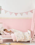 Child sleeping in toddler bed with pink floral duvet set and coordinating tiny tails nursery bunting draped across the wall.