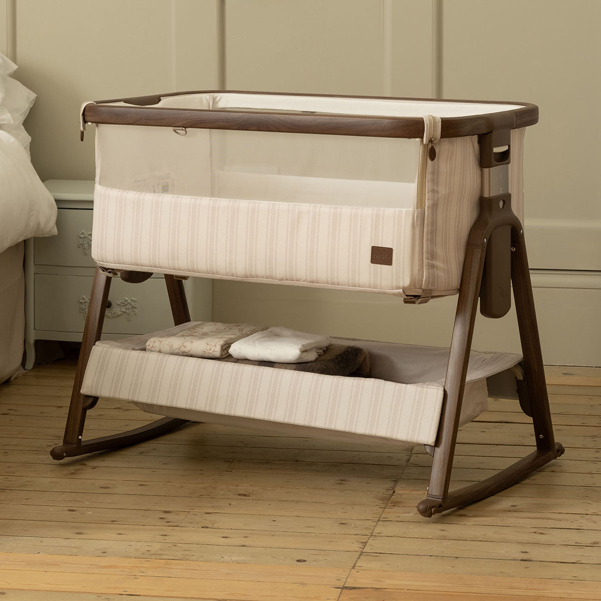 A baby bedside crib with a cream and beige striped fabric and a dark brown wooden frame, shown in a nursery setting on a light wood floor.