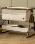 A baby bedside crib with a cream and beige striped fabric and a dark brown wooden frame, shown in a nursery setting on a light wood floor.