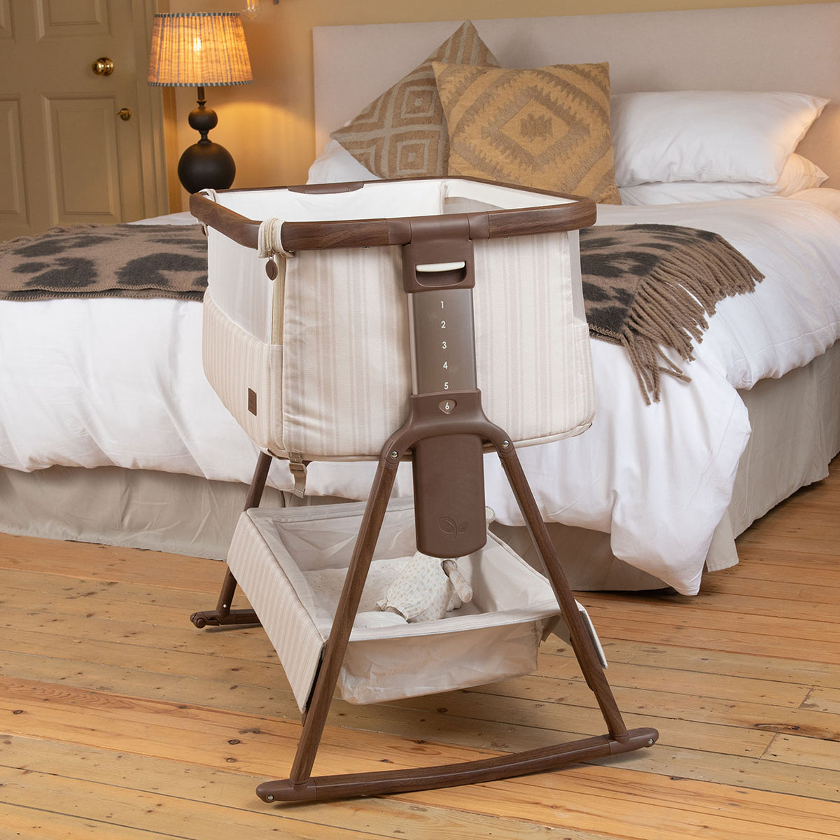 A baby bedside crib on a wooden rocking stand is placed in front of a neatly made bed in a bedroom. The crib has cream-colored fabric panels with a brown wooden frame and trim, and a matching fabric storage basket underneath.