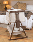 A baby bedside crib on a wooden rocking stand is placed in front of a neatly made bed in a bedroom. The crib has cream-colored fabric panels with a brown wooden frame and trim, and a matching fabric storage basket underneath.