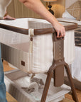 A person is shown from the side adjusting the wooden frame of a bedside crib in a bedroom setting. The crib has a cream and beige striped fabric lining, a dark brown wooden top frame, and a brown plastic height-adjustment handle with numbered slots.
