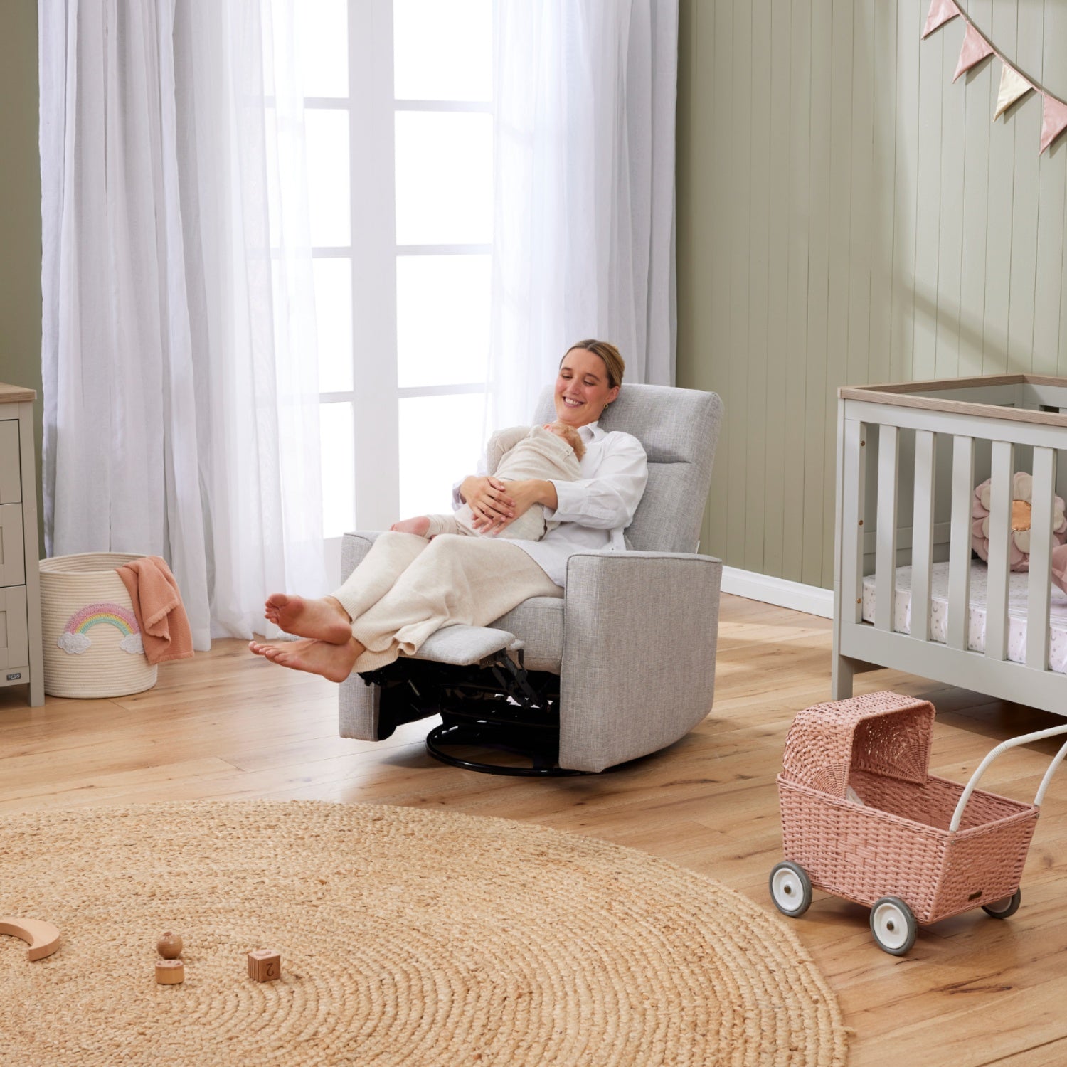 A woman is sitting in the Paige recliner chair in a nursery with a cot and toys around. 
