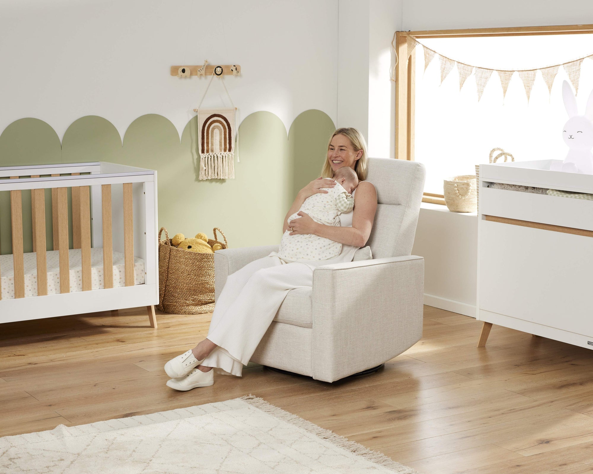 Woman in a white dress sitting in a chair in a nursery with a crib and dresser.