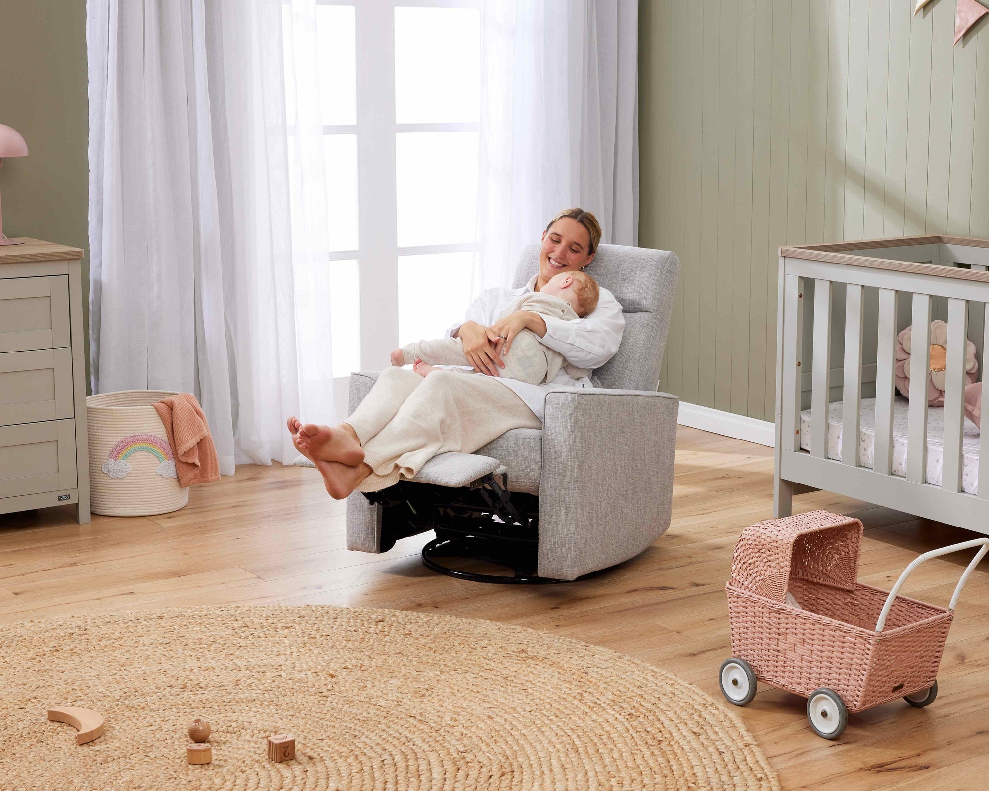 A woman sitting in a recliner chair holding a baby in a nursery with a cot and toys.