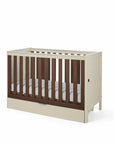 An angle view of the Riley cot bed in cashmere and rich walnut, featuring a drawer beneath it in a white background.