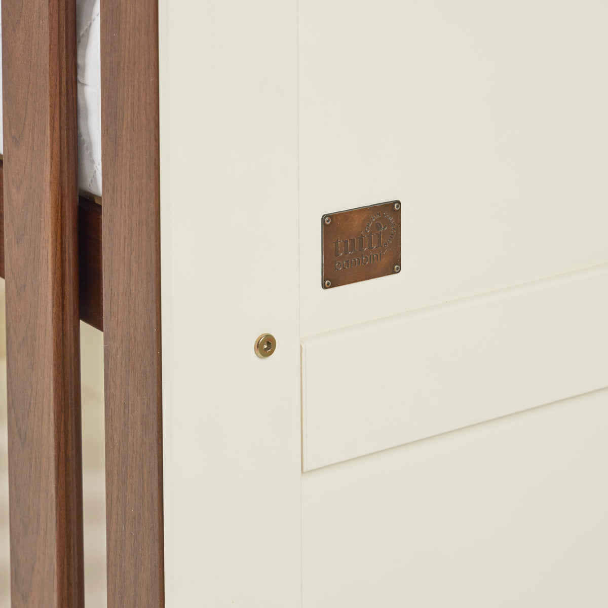 A close up view of the Riley cot bed in cashmere and rich walnut, featuring the Tutti Bambini Origin Collection nameplate screwed on the side of the bed.