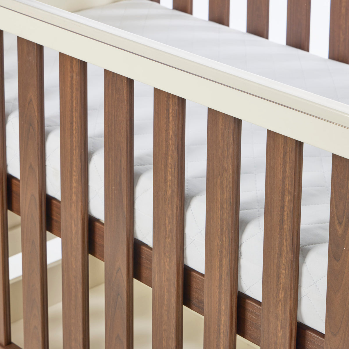 A close-up of the Riley cot bed with vertical slats and a white mattress inside, showing the rich walnut wood grain and cashmere frame details.