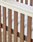 A close-up of the Riley cot bed with vertical slats and a white mattress inside, showing the rich walnut wood grain and cashmere frame details.