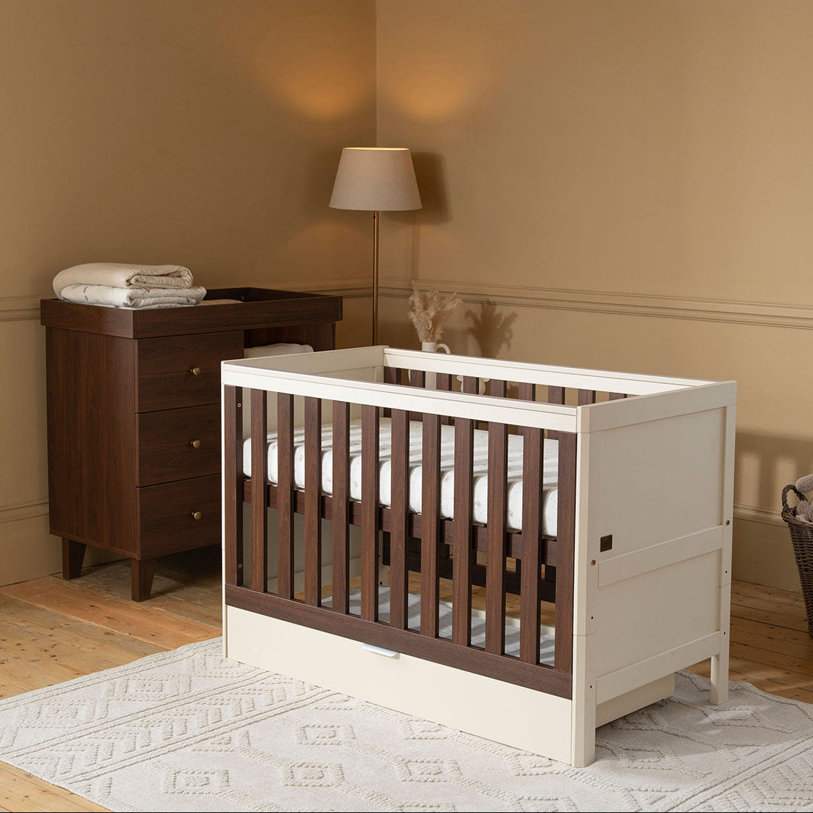 A white and dark brown wooden baby mini cot bed in a styled nursery setting with a matching dark brown changing table, a floor lamp, and beige walls.