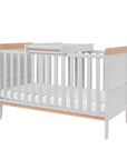 Rio cot bed in dove grey and oak shown from a side angle with removable cot top changer in place, highlighting practical changing functionality.