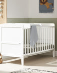 Side view of the Rio Cot Bed in white with cot top changer in a nursery room setting, showcasing slatted sides, solid panel ends, and a practical changer attachment for added functionality.