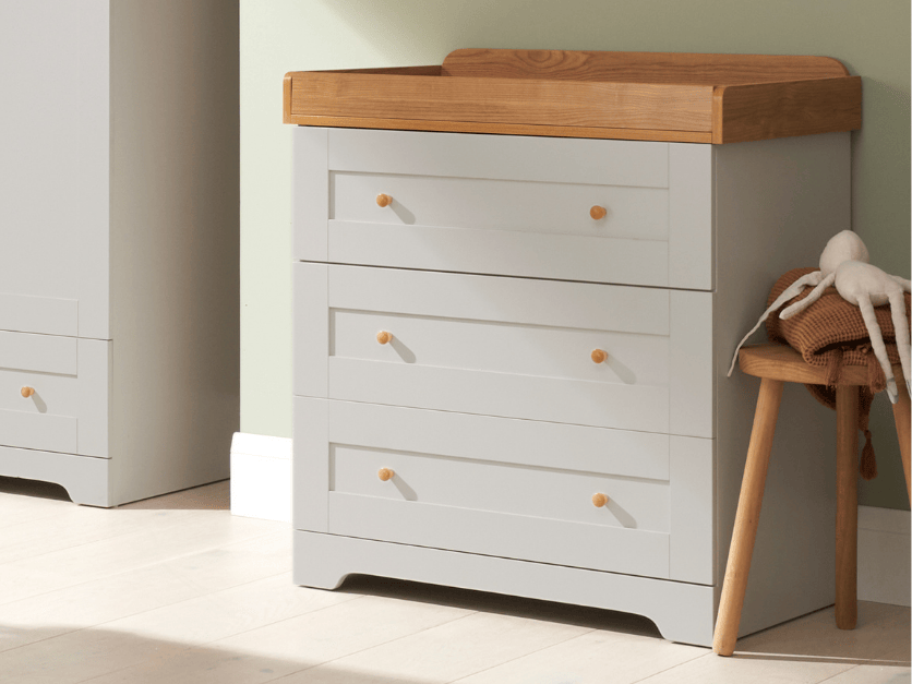 Angled lifestyle view of the Rio chest changer in dove grey with oak top, showcasing warm wood tones and classic detailing