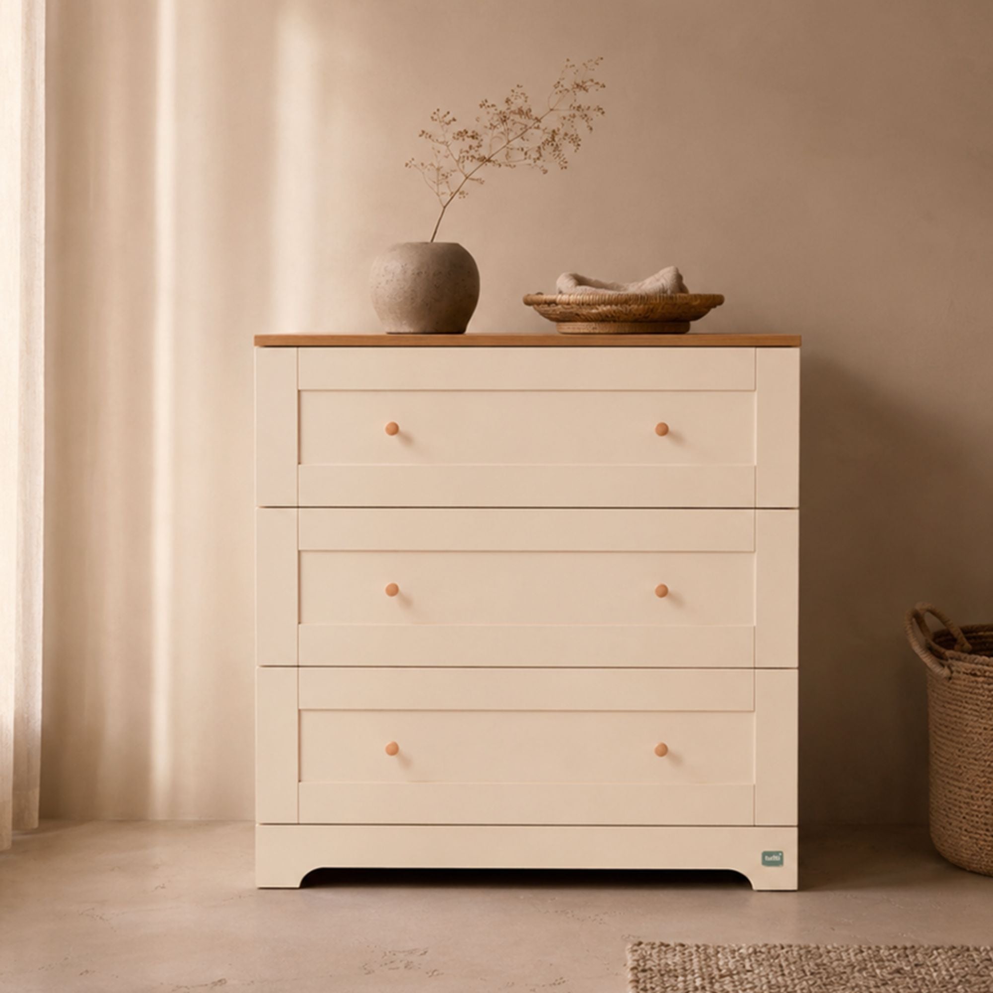A three-drawer cream chest of drawerswith a light wood top and small round wooden knobs, styled in a warm neutral room with a vase and dried stems on top.