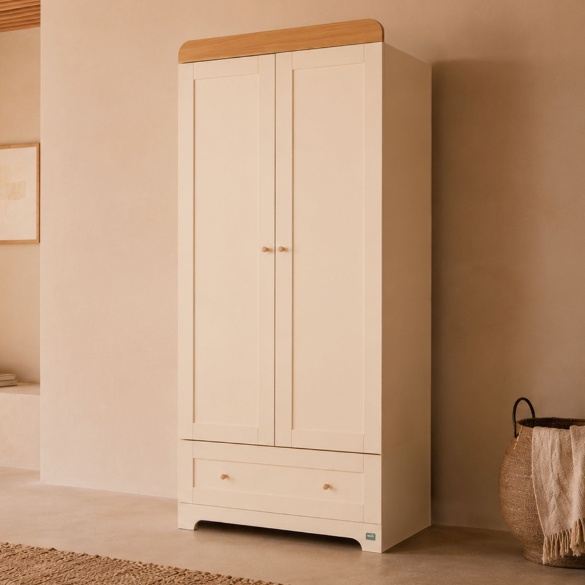 A tall cream-colored double-door wardrobe or armoire with a natural wood top and small drawer at the bottom, shown in a warm neutral-toned room setting.
